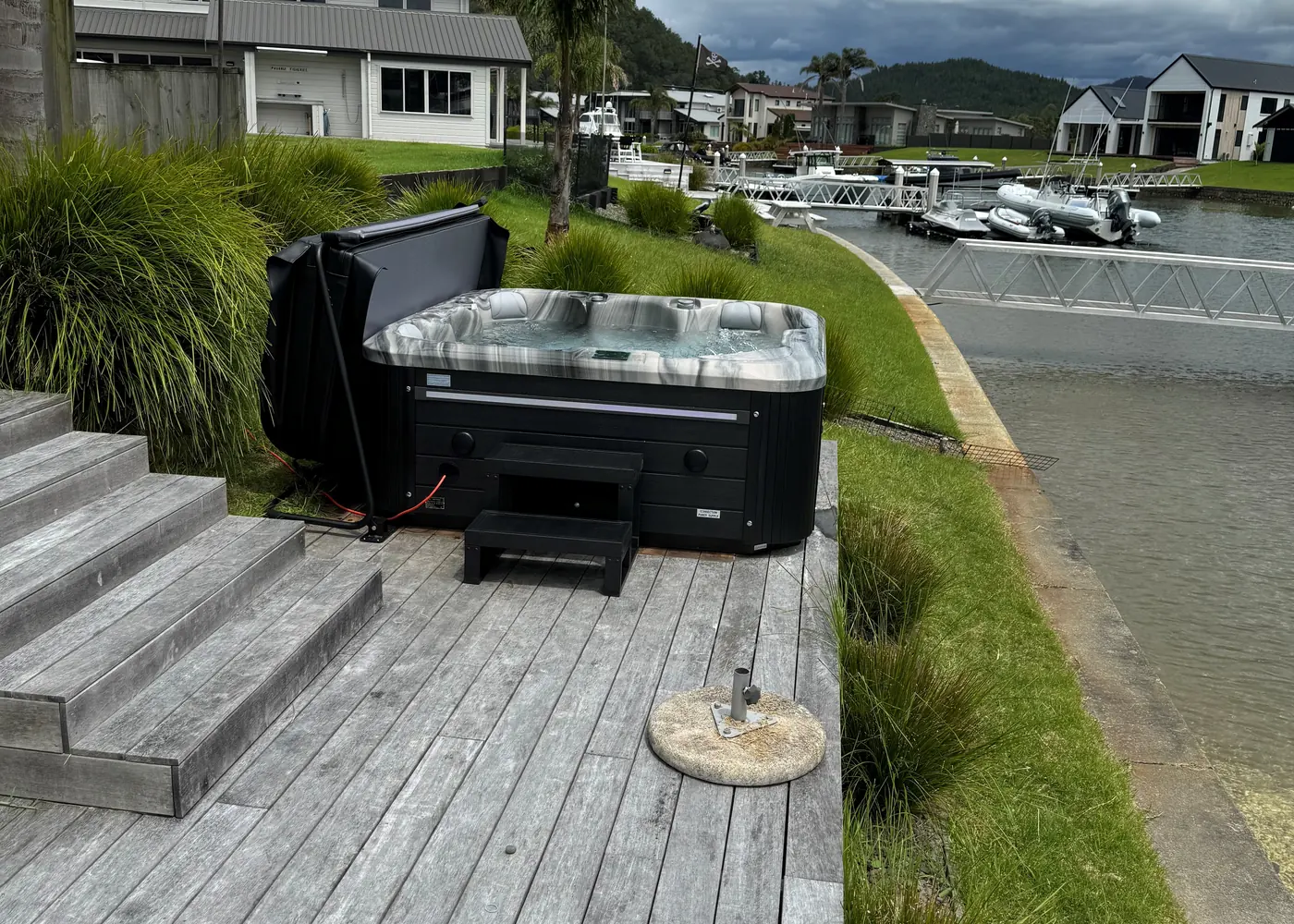 A spa pool on a timber deck beside the water, cover propped open